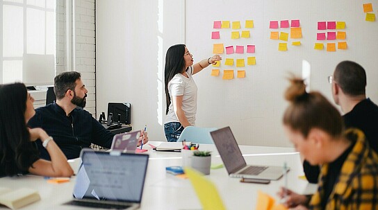 Frau steht erklärend an Wand, an der mehrere Post-its befestigt sind.