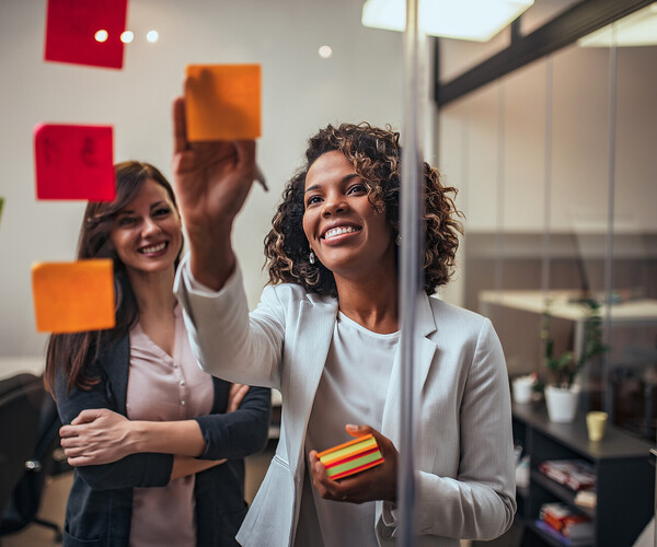 Creative businesswoman writing on sticky notes on a glass wall, female colleague looking. Frau klebt Klebezettel and Glaswand und unterhält sich mit anderer Frau