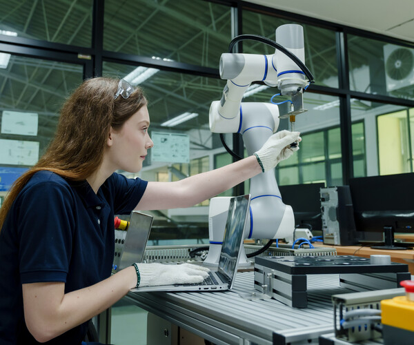 Junge Frau richtet in einem Labor einen Roboterarm ein und arbeitet dabei an einem Laptop.