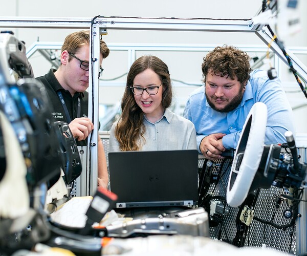 drei Arbeiter besprechen etwas an einem Laptop, der auf dem Sitz eines unfertigen Autos steht