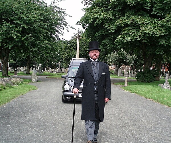 Bestatter mit dunkler Kleidung und Zylinder läuft Friedhof entlang, gefolgt von einem Auto