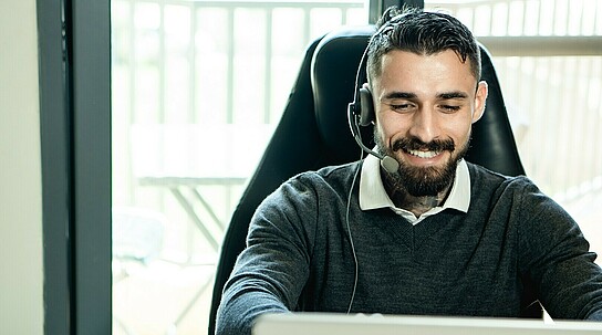 Man lächelt während er auf seinen PC guckt und ein Headset trägt