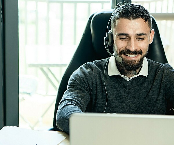 Man lächelt während er auf seinen PC guckt und ein Headset trägt