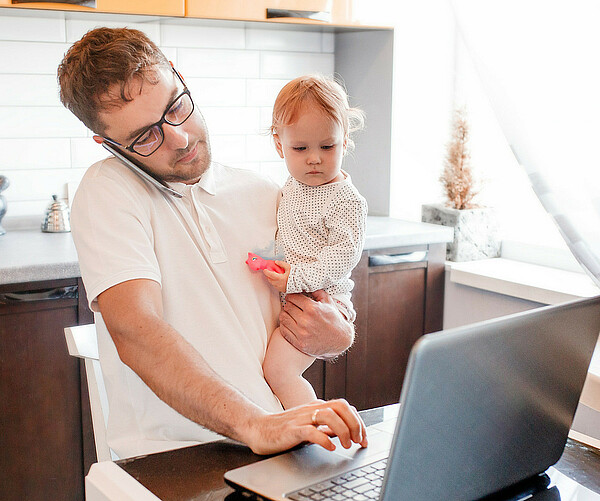 Mann arbeitet Zuhause an seinem Laptop, während er ein Baby im Arm hält und telefoniert