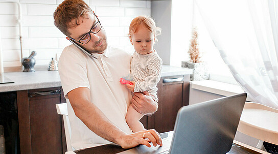 Mann arbeitet Zuhause an seinem Laptop, während er ein Baby im Arm hält und telefoniert