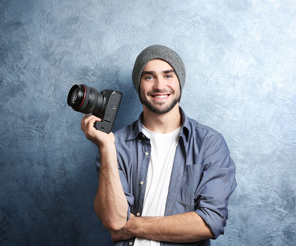 Handsome young photographer near grey textured wall Fotograf hält Kamera hoch und lächelt in die Kamera