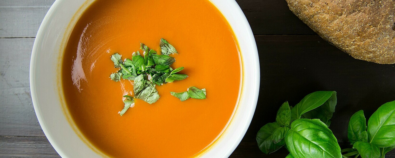 Tomatensuppe in einem Teller mit Basilikumblättern. Auf dem Tisch mehr Basilikum und dunkles Brot.