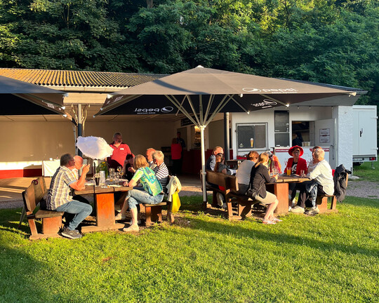 Impressionen der IHK-Sommertour 2025 in Podelwitz: Menschen sitzen am Holztisch draußen. Sie essen und reden.
