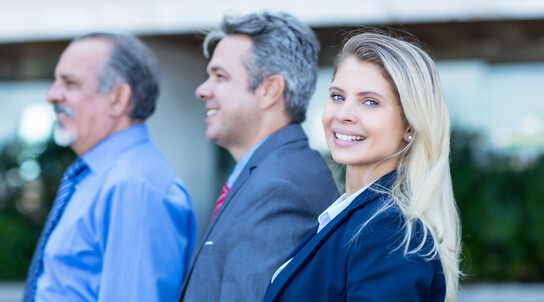 eine Frau steht vor zwei älteren Herren, alle im Business-Look