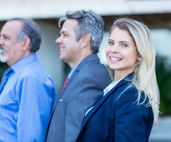 eine Frau steht vor zwei älteren Herren, alle im Business-Look