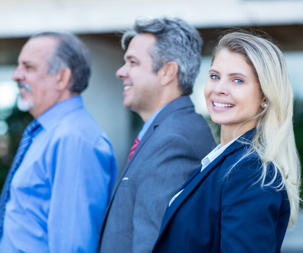 eine Frau steht vor zwei älteren Herren, alle im Business-Look