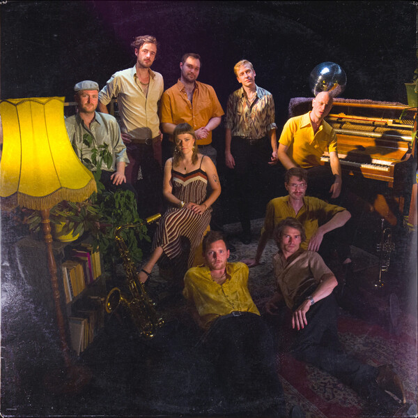 Neun Personen posieren in einem gemütlich eingerichteten Raum mit Stehlampe, Pflanzen, Saxofon und Klavier. Warmes Licht und eine Spiegelkugel verleihen dem Gruppenfoto eine Retro-Atmosphäre.