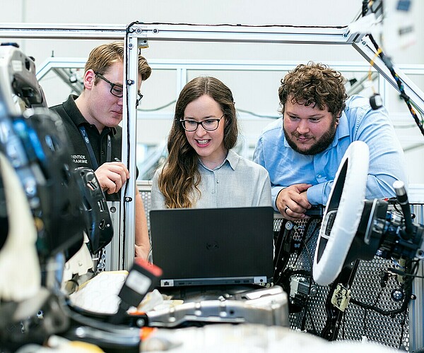 drei Arbeiter besprechen etwas an einem Laptop, der auf dem Sitz eines unfertigen Autos steht