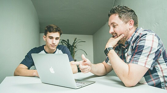Ein älterer Mann erklärt einem jüngeren Mann etwas an einem aufgeklappten Laptop