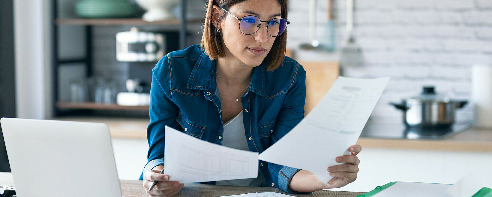 Pretty young business woman working with computer while consulting some invoices and documents in the kitchen at home. Symbolbild für E-Rechnungen, Frau mit Papierrechnung in der Hand sitzt am Laptop.