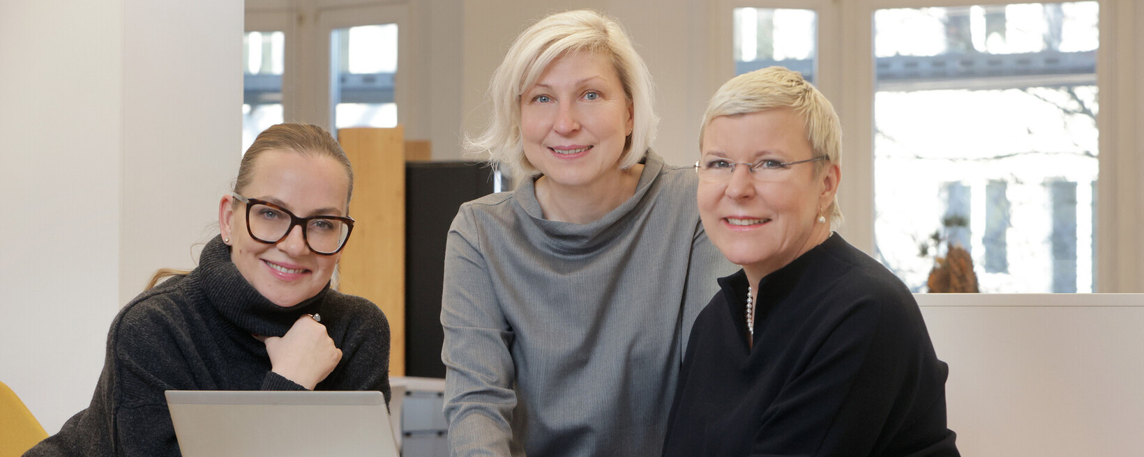 Drei Frauen sitzen lächelnd an einem Tisch mit Laptop und schauen in die Kamera.