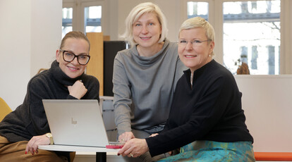 Drei Frauen sitzen lächelnd an einem Tisch mit Laptop und schauen in die Kamera.
