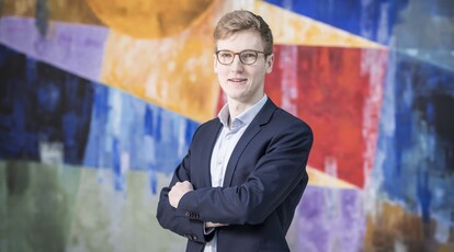 Porträt von Thorben Petri - junger Mann mit Brille und Anzug, der mit verschränkten Armen vor einem farbenfrohen, abstrakten Hintergrund steht.