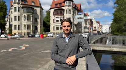 Jakob Ehrlich steht draußen in Leipzig, im Hintergrund sieht man die Straße und zwei alte Häuser sowie eine Brücke.