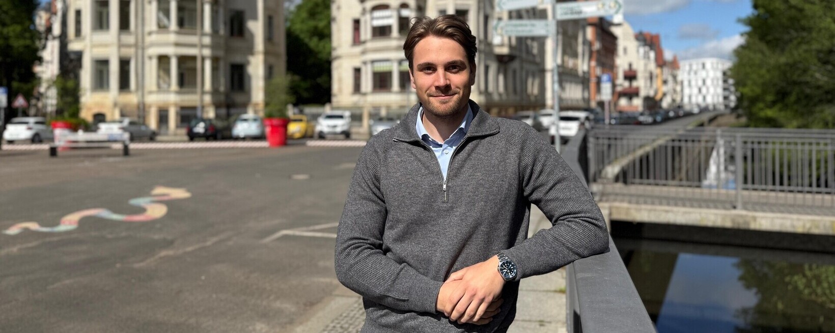 Jacob Ehrlich, KI-Experte (eigenes Bild) Jakob Ehrlich steht draußen in Leipzig, im Hintergrund sieht man die Straße und zwei alte Häuser sowie eine Brücke.